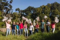 Retrato de un feliz grupo multiétnico de voluntarios de conservación limpiando el río en el campo, sosteniendo bolsas de basura en el aire. Ecología y responsabilidad social en el medio rural. - foto de stock