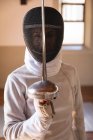 Retrato de esportista caucasiano vestindo roupa de esgrima protetora durante uma sessão de treinamento de esgrima, olhando para a câmera e sorrindo, segurando um epee em posição defensiva. Treinamento de esgrimistas em um ginásio. — Fotografia de Stock