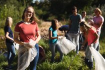 Портрет белой женщины-волонтёра, очищающей реку в сельской местности, её друзья собирают мусор на заднем плане. Экология и социальная ответственность в сельской местности. — стоковое фото