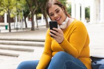 Curvy femme caucasienne dans les rues de la ville pendant la journée, assis sur des marches, souriant et utilisant son smartphone portant des écouteurs avec un bâtiment historique en arrière-plan — Photo de stock