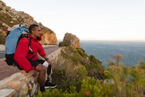 Athlète masculin en forme, handicapé, de race mixte avec une jambe prothétique, profitant de son temps sur un voyage à la montagne, randonnée, assis sur un mur sur la route par la mer. Mode de vie actif avec handicap. — Photo de stock