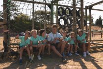 Группа кавказских мальчиков и девочек, сидящих с кавказским тренером по фитнесу, отдыхающих, пьющих бутылки воды и разговаривающих в лагере в солнечный день — стоковое фото