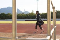 Athlète masculin handicapé de race mixte dans un stade de sport de plein air, marchant avec un sac de sport et une bouteille d'eau sur une piste de course portant des lames de course. Handicap athlétisme entraînement sportif. — Photo de stock
