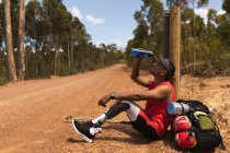 A fit, disabled mixed race male athlete with prosthetic leg, enjoying his time on a trip, hiking, sitting on a dirt road in a forest, pouring water on himself. Active lifestyle with disability. — Stock Photo