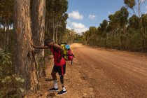 Athlète masculin de race mixte en forme et handicapé avec une jambe prothétique, profitant de son temps lors d'un voyage, d'une randonnée, appuyé sur un arbre sur un chemin de terre dans une forêt. Mode de vie actif avec handicap. — Photo de stock
