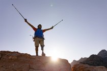 Fit, disabled mixed race male athlete with prosthetic leg, enjoying his time on a trip to the mountains, hiking on the rocks with his arms above his head with sticks. Active lifestyle with disability. — Stock Photo