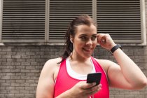Улыбающаяся кривая белая женщина с длинными темными волосами в спортивной одежде, тренирующаяся в городе, пользующаяся своим смартфоном с наушниками, кирпичная стена на заднем плане — стоковое фото