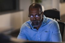 Uomo d'affari afroamericano che lavora fino a tardi la sera in un ufficio moderno, seduto a una scrivania, utilizzando un computer desktop. — Foto stock