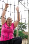 Femme de race mixte portant un t-shirt rose lors d'une séance d'entraînement au camp d'entraînement, faisant de l'exercice, grimpant sur des filets au-dessus d'un cadre d'escalade. Exercice de groupe en plein air, défi sain amusant. — Photo de stock