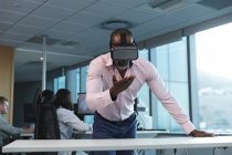 Afroamerikanischer Geschäftsmann arbeitet spät abends in einem modernen Büro, lehnt an einem Schreibtisch, trägt ein Vr-Headset, blickt auf einen virtuellen Bildschirm, im Hintergrund seine Kollegen.. — Stockfoto