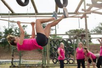 Fit femme caucasienne portant t-shirt rose lors d'une session d'entraînement du camp d'entraînement, l'exercice, accroché à une corde avec ses mains et ses pieds, groupe la motivant. Exercice de groupe en plein air, défi sain amusant. — Photo de stock