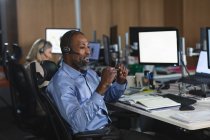 Hombre de negocios afroamericano que trabaja hasta tarde en la noche en una oficina moderna, sentado en un escritorio, con auriculares telefónicos y hablando. - foto de stock