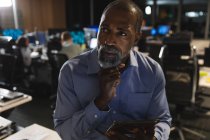 Homme d'affaires afro-américain travaillant tard dans la soirée dans un bureau moderne, assis sur un bureau, utilisant une tablette informatique, tenant son menton et pensant. — Photo de stock