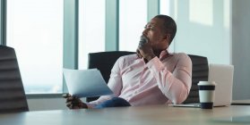 Empresario afroamericano que trabaja hasta tarde en una oficina moderna, sentado en un escritorio, sosteniendo un pedazo de papel y pensando:. - foto de stock