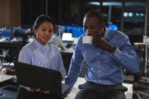 Femme d'affaires asiatique et homme d'affaires afro-américain travaillant tard dans la soirée dans un bureau moderne, assis sur un bureau, utilisant un ordinateur portable, l'homme tenant une tasse et buvant. — Photo de stock