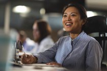 Азійська комерсантка працює пізно ввечері в сучасному офісі, сидячи за столом, роблячи нотатки і посміхаючись, з колегами на задньому плані.. — стокове фото