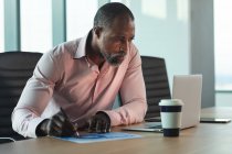 Homme d'affaires afro-américain travaillant tard dans la soirée dans un bureau moderne, assis à un bureau et utilisant un ordinateur portable, prenant des notes. — Photo de stock