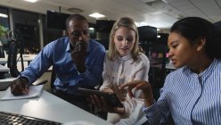Groupe multiethnique de collègues travaillant tard dans la soirée dans un bureau moderne, assis à un bureau, utilisant une tablette et discutant de leur travail. — Photo de stock