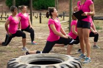 Groupe multi-ethnique de femmes portant toutes des t-shirts roses lors d'une séance d'entraînement au camp d'entraînement, faisant de l'exercice, étirant les jambes et le canapé les motivant. Exercice de groupe en plein air, défi sain amusant. — Photo de stock