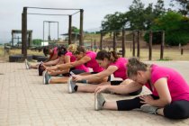 Groupe multi-ethnique de femmes portant toutes des t-shirts roses lors d'une séance d'entraînement dans un camp d'entraînement, faisant de l'exercice, étirant les jambes, s'asseyant sur le sol. Exercice de groupe en plein air, défi sain amusant. — Photo de stock