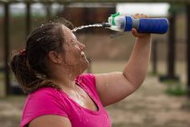 Une femme de race mixte portant un t-shirt rose lors d'une séance d'entraînement au camp d'entraînement, faisant de l'exercice, faisant une pause, versant de l'eau sur son visage. Exercice de groupe en plein air, défi sain amusant. — Photo de stock