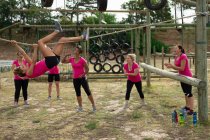 Fit mulher caucasiana vestindo camiseta rosa em uma sessão de treinamento do acampamento de inicialização, exercitando, pendurado em uma corda com as mãos e os pés, grupo motivando-a. Exercício de grupo ao ar livre, desafio saudável divertido. — Fotografia de Stock