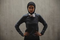 Portrait de femme de race mixte en hijab, écouteurs et vêtements de sport faisant de l'exercice à l'extérieur dans la ville, regardant la caméra. Exercice mode de vie urbain. — Photo de stock