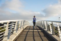 Встановіть змішану жінку в хіджабі і спортивному одязі, займаючись на відкритому повітрі в місті в сонячний день, бігаючи на пішохідному мосту. Міський спосіб життя вправи . — стокове фото