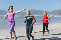 Gruppo di amiche caucasiche che si esercitano su una spiaggia in una giornata di sole, corrono sulla riva del mare e sorridono. — Foto stock