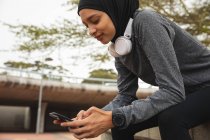 Fit mulher de raça mista vestindo hijab e sportswear exercitando ao ar livre na cidade, sentado fazendo uma pausa usando seu smartphone no parque urbano. Exercício urbano. — Fotografia de Stock