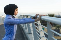Подходящая смешанная расовая женщина в хиджабе и спортивной одежде занимается спортом на открытом воздухе в солнечный день, растягиваясь на пешеходном мосту. Упражнения городского образа жизни. — стоковое фото