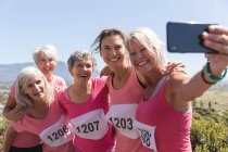 Group of Caucasian female friends enjoying exercising on a sunny day, celebrating after running race, wearing numbers and smiling, taking a photo with smartphone. — Stock Photo