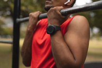 In der Mitte ein Mann in Sportkleidung, der in einem Park im Outdoor-Fitnessstudio mit drahtlosen Kopfhörern und Smartwatch trainiert und Klimmzüge macht. Fitness stärkt gesunden Lebensstil. — Stockfoto