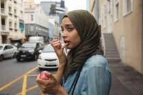 Mischlingshündin mit Hidschab unterwegs in der Stadt, die auf der Straße steht und Lippenbalsam aufträgt, während der Straßenverkehr hinter ihr steht. Pendler moderner Lebensstil. — Stockfoto