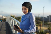 Ajuste mujer de raza mixta que usa hijab y ropa deportiva haciendo ejercicio al aire libre en la ciudad en un día soleado, tomando un descanso durante el entrenamiento usando teléfonos inteligentes y auriculares en una pasarela. Ejercicio urbano. - foto de stock
