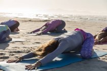 Grupo de amigas caucásicas disfrutando haciendo ejercicio en una playa en un día soleado, practicando yoga y sentadas en posición de yoga. - foto de stock