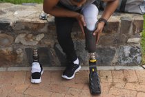 Partie basse d'un homme handicapé de race mixte avec une jambe prothétique travaillant dans un parc urbain, assis sur un mur et ajustant une lame de course. Fitness handicap mode de vie sain. — Photo de stock