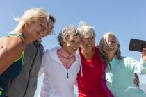 Gruppo di amiche caucasiche che si divertono ad allenarsi su una spiaggia in una giornata di sole, scattando foto con uno smartphone, sorridendo e abbracciando. — Foto stock