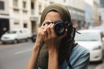 Mulher de raça mista vestindo hijab passear para fora e sobre em movimento na cidade, sorrindo segurando câmera digital tirar fotos. Turismo sightseeing estilo de vida moderno. — Fotografia de Stock
