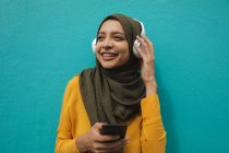 Femme de race mixte portant hijab et pull jaune sur et sur la route dans la ville, souriant tenant smartphone avec casque sans fil sur. Commuter style de vie moderne. — Photo de stock