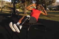 Homem de raça mista deficiente com uma perna protética vestindo roupas esportivas, trabalhando em um parque, ouvindo música em fones de ouvido, fazendo crunches. Incapacidade física estilo de vida saudável. — Fotografia de Stock