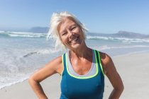 Портрет пожилой кавказки, занимающейся спортом на пляже в солнечный день, улыбающейся, стоящей и смотрящей в камеру с морем на заднем плане. — стоковое фото