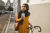 Femme de course mixte portant hijab et pull jaune sur et sur la route dans la ville, tenant café à emporter à pied avec vélo. Commuter style de vie moderne. — Photo de stock