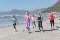 Gruppo di amiche caucasiche che si esercitano su una spiaggia in una giornata di sole, corrono sulla riva del mare e sorridono. — Foto stock