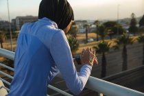 Rear view of fit mixed race woman wearing hijab and sportswear exercising outdoors in the city on a sunny day, checking her smartwatch on a footbridge. Urban lifestyle exercise. — Stock Photo