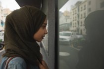 Mujer de raza mixta vistiendo hijab fuera y sobre la marcha en la ciudad, de pie en la calle mirando por el escaparate. Commuter estilo de vida moderno. - foto de stock
