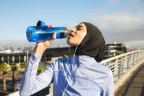 Fit gemischte Rennläuferin in Hijab und Sportbekleidung, die an einem sonnigen Tag im Freien in der Stadt trainiert und aus einer Wasserflasche trinkt, während sie auf einer Fußgängerbrücke Kopfhörer trägt. Bewegung im städtischen Lebensstil. — Stockfoto