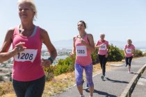 Gruppo di amiche caucasiche che si divertono ad allenarsi in una giornata di sole, a correre, a indossare numeri e abbigliamento sportivo rosa, a sorridere. — Foto stock