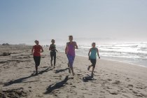 Groupe d'amies caucasiennes qui aiment faire de l'exercice sur une plage par une journée ensoleillée, courir sur le bord de la mer et sourire. — Photo de stock
