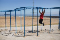Disabled mixed race man with a prosthetic leg and running blade exercising at an outdoor gym by the coast, working out on the monkey bars. Fitness disability healthy lifestyle. — Stock Photo